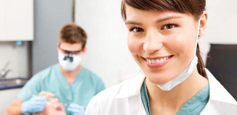 A female dental assistant smiles as a male dental assistant works on a patient in the background